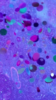 Dikey video. Parıldayan sıvı. Konfeti döküldü. Dekoratif kıvılcımları olan su mor buz yüzeyinden aşağı akar. Bulanık mürekkep neon efektli renkli arkaplan.