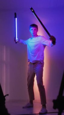 Dikey video. Fütürist poz. Neon eğitimi. LED tüpleri ışın kılıcı gibi sallayan adam eğleniyor. Profesyonel gece stüdyosunda video çekimi.