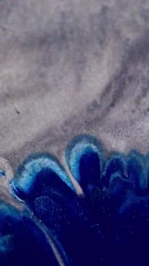 Dikey video. Mürekkep lekesi. Parıltı karışımı. Parlak mavi lekeli sıvı gümüş gri parıldayan parçacıkların yüzeyine yayılmış. Kontrast rengi soyut odaklanmış arkaplan.