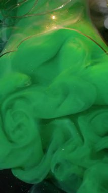 Dikey video. Sanat buharı. Boya bulutu. Suyun siyah arka planında kırık bir ampulden sisli yeşil duman çıkıyor. Yaratıcı sıvı soyut yüzey.