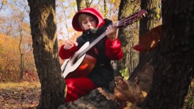 Sonbaharda ağaçların arasında bir çocuk bir kütüğe oturur ve neşeyle gitar çalar.