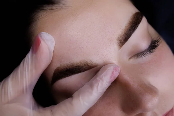 Permanent makeup of the eyebrows, the fingers of the master stretch the eyebrow of the model to show the tattoo of the eyebrows