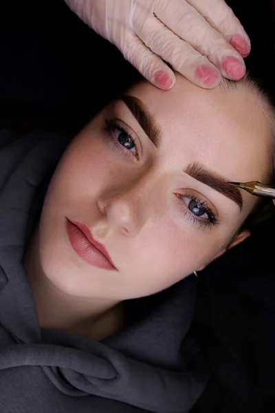 Permanent eyebrow makeup, the master holds a tattoo machine near the model\'s eyebrows showing the result after tattooing