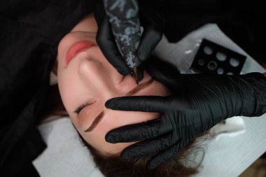 Usta, modelin kaşlarını parmaklarıyla tutuyor ve kaşlarını kalıcı olarak uyguluyor. Kaş kalıcı makyaj kozmetik prosedürü.