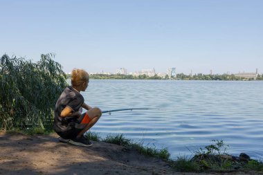 Bir nehrin kıyısında, bir çocuk bir şamandırayı izlerken balık tutuyor. Yazın nehirde spor balıkçılığı.