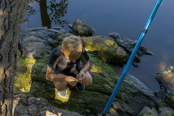 A boy sitting on rocks by the river equips the hook with bait. Sport fishing on the river in summer.