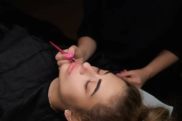 The master with a brush applies a toning paste to the lip contour Delicate permanent lip makeup for blondes
