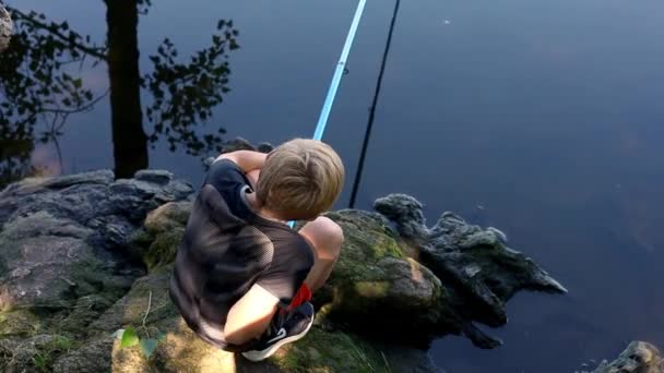 釣り糸を投げる岩の上の岸に座っている十代の少年 — ストック動画