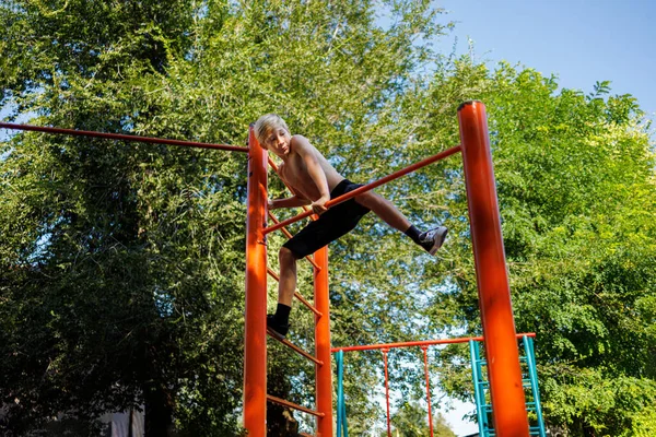 A boy athlete climbs a horizontal bar to perform a formal element. Street workout on a horizontal bar in the school park.