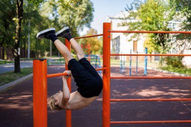 Bir genç bacaklarını yatay çubuğa atarak egzersiz yapıyor. Okul parkındaki yatay bir barda sokak çalışması..