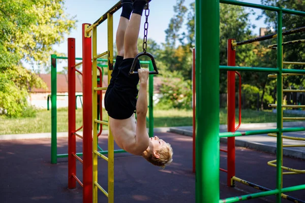 stock image The teenager hangs upside down, holding on to the handles on the chains. Street workout on a horizontal bar in the school park.