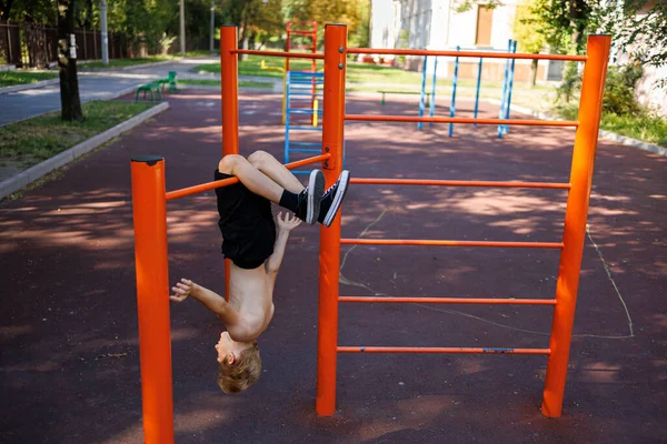 a teenager of an athletic build hangs head down, clinging to the horizontal bar with his feet. Street workout on a horizontal bar in the school park.