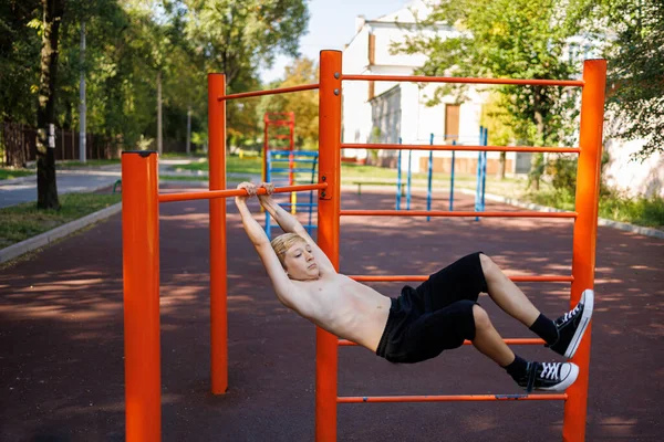 An athletic teenager sways in different directions, hanging on the horizontal bar. Street workout on a horizontal bar in the school park.