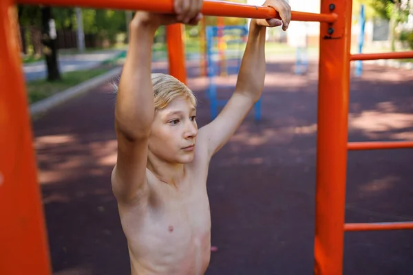 A teenager with an athletic development of musculature holds on to the horizontal bar. Street workout on a horizontal bar in the school park.