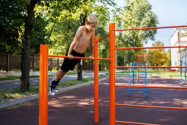 ティーンエイジャーは 学校の公園の水平バーでバーストリートワークアウトの要素の実行中に拡張された腕に彼の体重を保持します — ストック写真