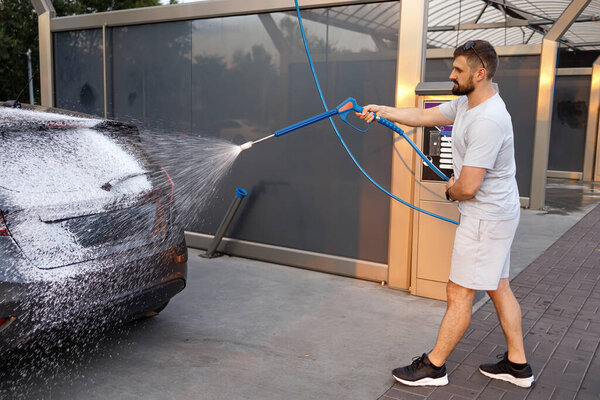 A man holds a foam sprayer from which foam flies out onto a car. A car at a self service car wash.