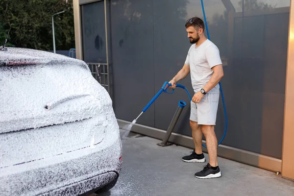 The rear of the car is covered with foam, the man in the background is applying foam to the car. A car at a self service car wash.