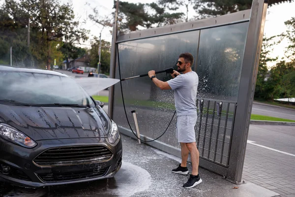 A man at the car wash points a water cannon at the windshield. A car at a self service car wash.