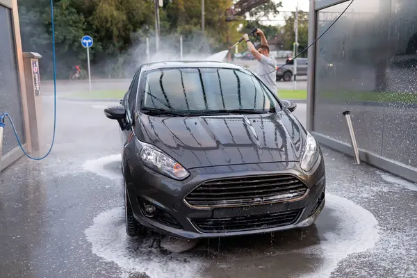 The front of a washed car, with a man washing the car in the background. A car at a self service car wash.