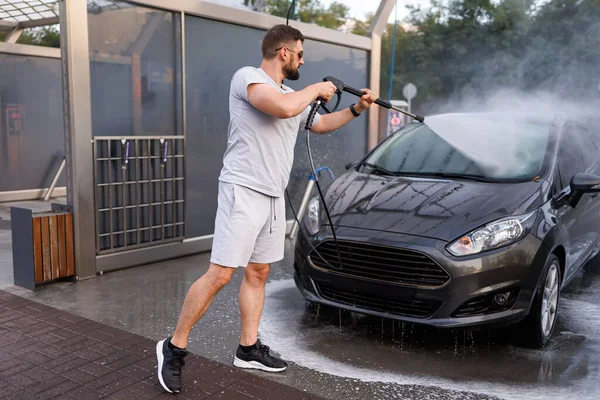 A man stands in front of the car and points a water cannon at it, washing the water off of it. A car at a self service car wash.