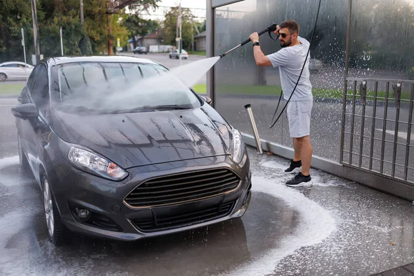 A man thoroughly washes the dirt off the car with a water cannon. A car at a self service car wash.
