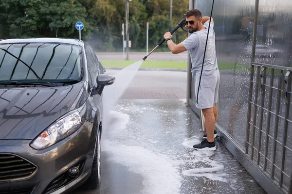 A man thoroughly washes the dirt off the car with a water cannon. A car at a self service car wash.