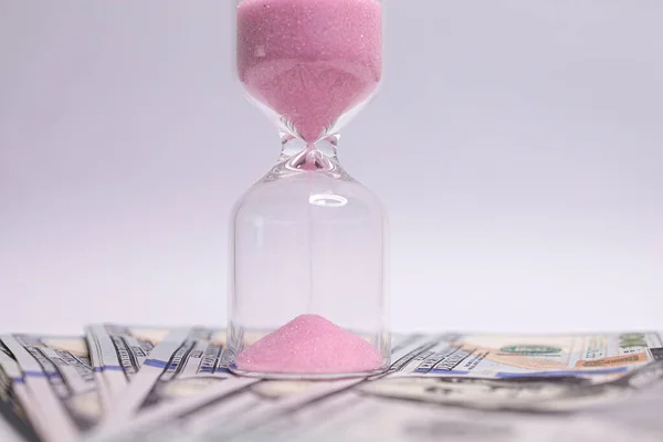 Close-up of the hourglass that stands on dollar bills. The hourglass is located near the dollar bills.