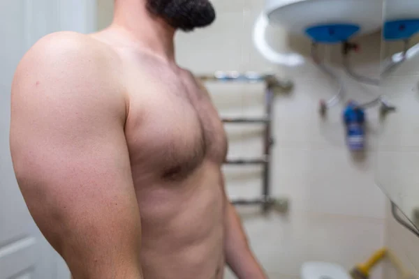 The body of a man standing in the bathroom. A man with an athletic figure in the bathroom.