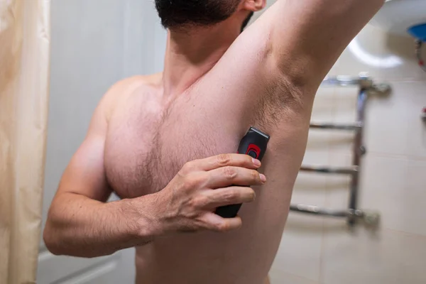 A man removes his armpit hair with an electric razor. A man with an athletic figure in the bathroom.