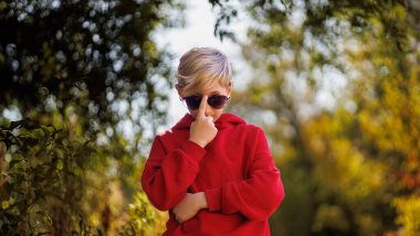 Çocuk güneş gözlüğünü parmağıyla düzeltir. Sonbaharda parkta kırmızı elbiseli bir çocuk..