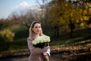Sonbahar parkında, bir iş kadını bir buket gülü kucaklıyor. Sonbaharda bir buket beyaz gülle ceketli şık bir kadın..