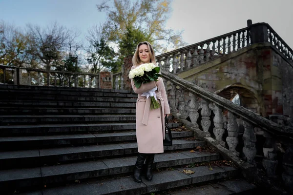 A beautiful woman with a bouquet in her hands stands near an ancient staircase. Stylish woman in a coat in autumn with a bouquet of white roses.