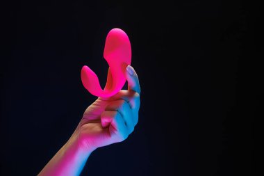 Kadınların elleri kırmızı renkli vibratörü parmaklarıyla nazikçe tutuyor. Siyah arka planda neon ışıkları olan bir seks oyuncağı klitoral vibratörü..