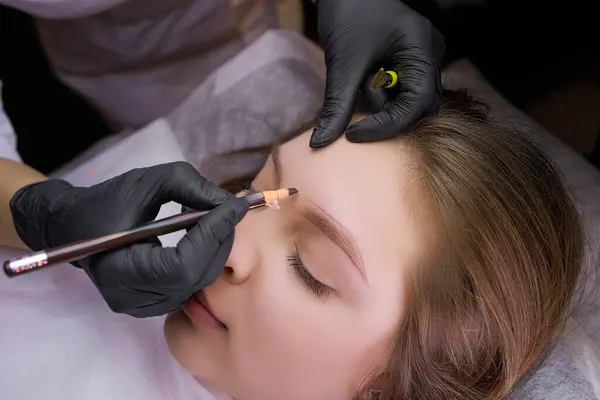 Kaş kalemi yardımıyla, usta kalıcı makyaj işleminden önce dış hatlara bir işaret koyar. PMU Prosedürü, Kalıcı Kaş Makyajı.