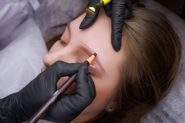 Kaş kalemi yardımıyla, usta kalıcı makyaj işleminden önce dış hatlara bir işaret koyar. PMU Prosedürü, Kalıcı Kaş Makyajı.