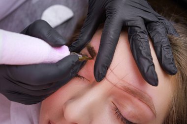 Usta dövme makinesinin ellerinde, kaşlarına kalıcı makyaj uyguluyor. PMU Prosedürü, Kalıcı Kaş Makyajı.