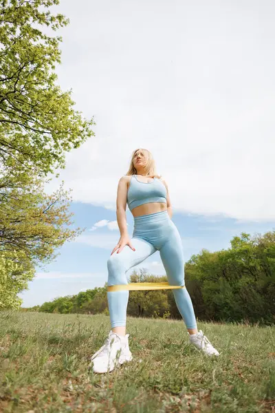 A girl exercises with a rubber band on her on her legs. Beautiful blonde Caucasian woman in blue tight tracksuit. Blonde girl at an outdoor training session