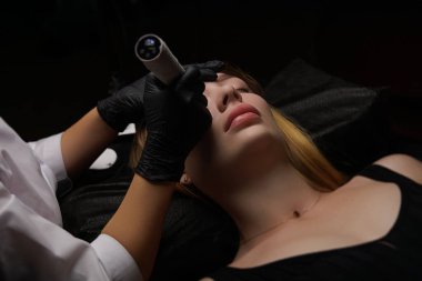 Müşteri koltukta rahatça otururken bir cilt bakımı uzmanı, hastanın kaşlarının durumunu iyileştirmek için bir makina uyguluyor. Kalıcı kaş makyajı, PMU için kahverengi pigmentli kaş dövmesi..