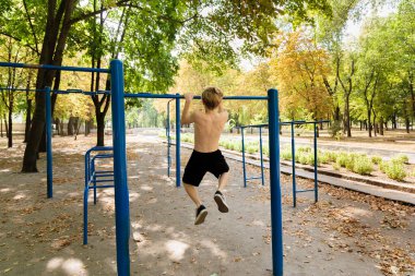 Parkta genç bir kız kendini barın üstüne çekerken arkadan bak. Parkta spor yapan genç bir çocuk. 