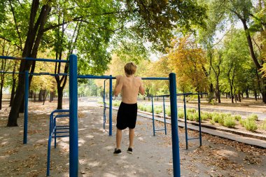 Genç bir çocuk barın üstünde barfiks çekiyor. Parkta spor yapan genç bir çocuk. 