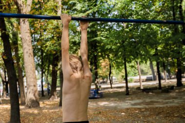 Atletik bir genç çocuk direğe asılmış ve aşağıya bakıyor. Parkta spor yapan genç bir çocuk. 