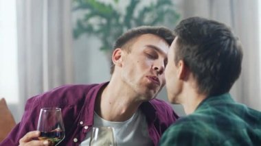Evde romantik bir randevu sırasında partnerini öpen ve şarap içen mutlu genç bir adamın portresi. Homoseksüel erkek çiftin birlikte vakit geçirmesini seviyorum. LGBT ilişki konsepti. 4K görüntü