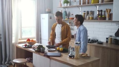 Happy young gay couple making breakfast together and talking before work day at the kitchen. Cheerful LGBT family spending pleasant morning making dishes together. High quality 4K footage