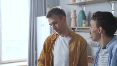 Happy morning of loving homosexual male preparing breakfast for his partner at the kitchen. Same-sex couple living their happy domestic life, cooking dishes together, kissing and smiling. LGBT concept