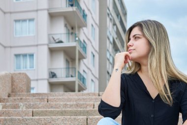 Plajda görülenlere benzer granit bir merdivende oturan Latin kız. Profilde sola bakıyor, arka planda binalar görülebilir..