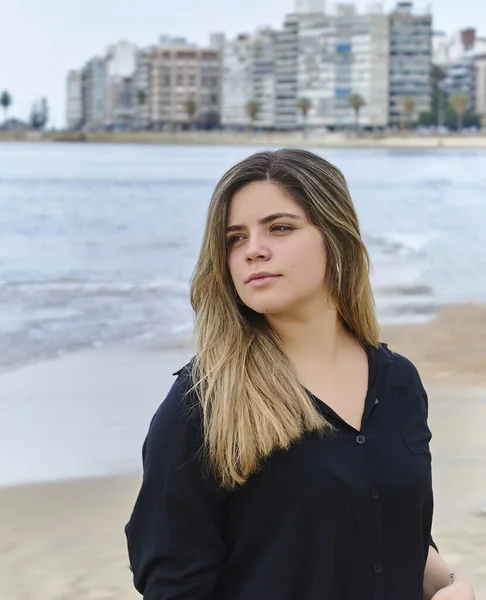 Sahilde arka planda binalar olan genç bir kız. Kumu görebilirsin. Kızın kendine güveni tam, denize bakıyor. Kız siyah bir gömlek giyiyor..