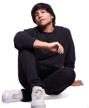 20 'li yaşlarda genç bir adam yerde bağdaş kurmuş oturuyor. Siyah bir sweatshirt ve beyaz kot pantolon giyiyor. Çocuk beyaz bir arka planda..