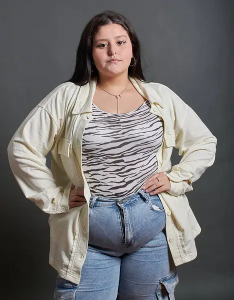 Genç Latin kız bir fotoğraf stüdyosunda poz veriyor, American shot. Genç kızın elleri belinde, kameraya bakıyor, tarafsız bir arkaplan..