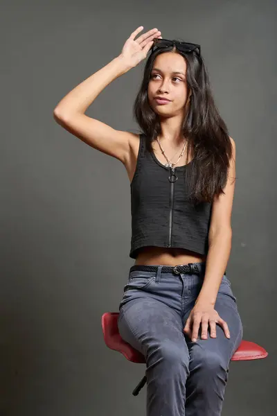 Kırmızı sandalyede oturan genç bir manken. Önünde fermuarı ve gri pantolonu olan siyah kolsuz bir bluz giyiyor. Sağ el kafasının üstüne kaldırılır ve güneş gözlüğü tutar..