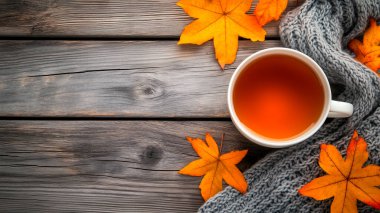 Sonbahar yapraklı bir fincan çay ve yukarıdan görünen ahşap bir masanın üzerinde yün bir eşarp. Sonbahar teması mevsimi ılık bir renk paletiyle anımsatıyor.